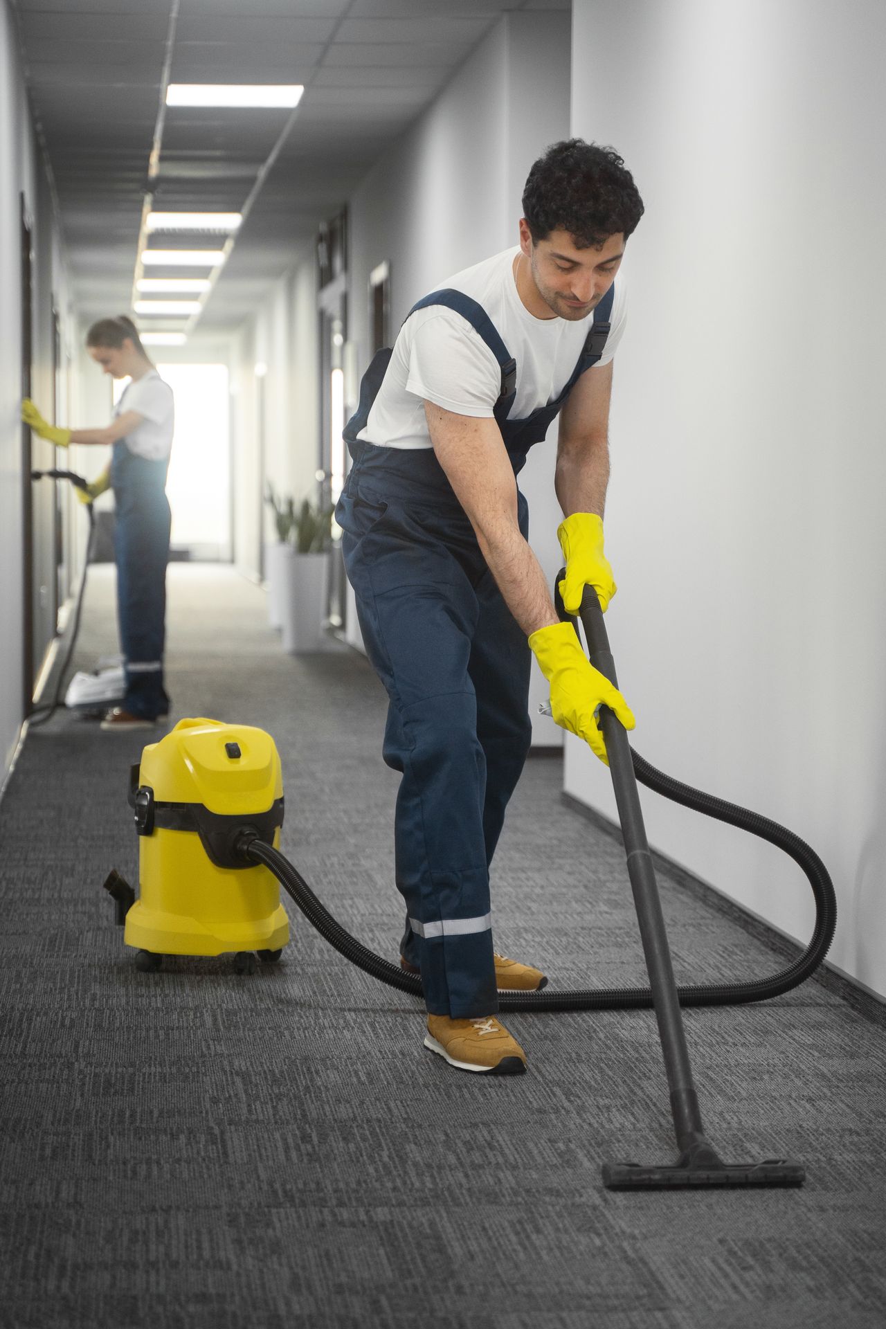 Professional cleaner vacuuming carpet in hallway with industrial cleaning equipment and safety gloves